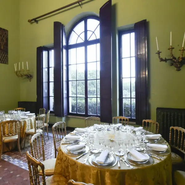 Sala interna con pareti verde salvia e alte finestre ad arco, tavoli rotondi apparecchiati con tovaglie dorate, cristalli e argenteria, appliques dorate e un ritratto a olio alla parete