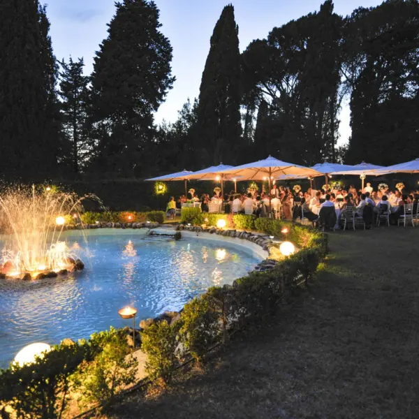 Ricevimento all’aperto al crepuscolo: un getto d’acqua illuminato al centro del laghetto, gli ospiti seduti ai tavoli sotto grandi ombrelloni bianchi, lampade sferiche accese lungo la siepe e fiaccole sul prato, cipressi e pini scuri contro il cielo blu notte