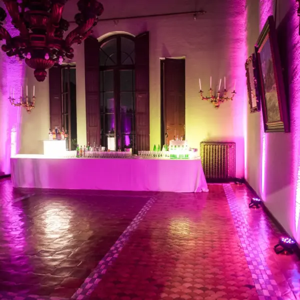 Sala interna illuminata di fucsia e ambra da fari a terra, un lampadario ligneo intagliato, alte finestre ad arco con scuri in legno, ritratti a olio e specchiere dorate alle pareti, un lungo banco bar con tovaglia bianca e bottiglie, pavimento in cotto a scaglie