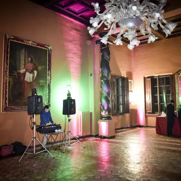 Salone con pareti color terracotta e soffitto a cassettoni, grande lampadario in vetro di Murano bianco, un ritratto a olio di cardinale in cornice dorata, una colonna tortile dipinta a tralci di fiori, un banco bar con tovaglia rossa e la consolle di un disc jockey, pavimento in cotto decorato