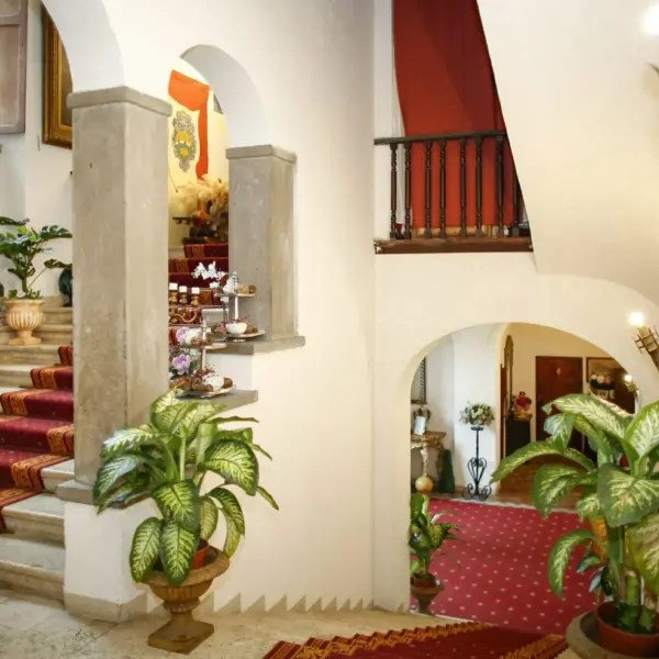 Scalone interno in pietra con passatoia rossa e oro, arcate in pietra chiara, un ballatoio ligneo con drappo rosso, stemmi araldici dipinti alla parete, torciere in ferro battuto, vasi di terracotta con piante e alzate con dolci e fiori sui gradini