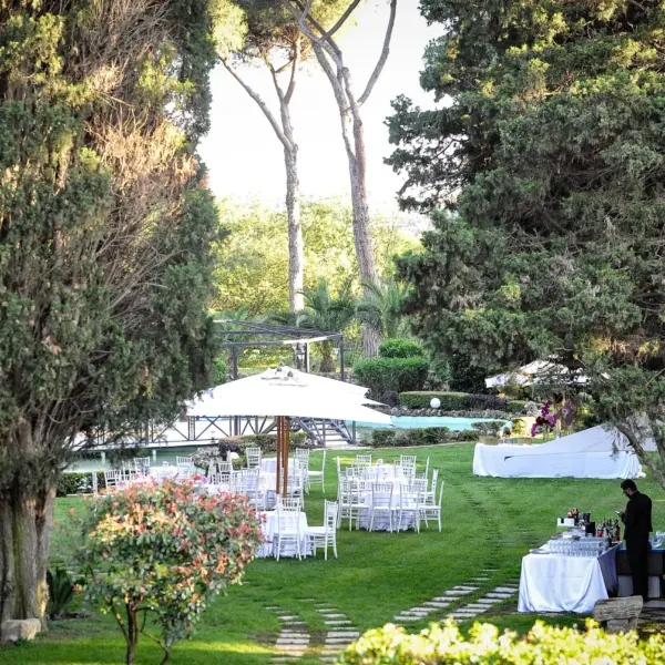Tavoli rotondi con tovaglie bianche e sedie chiavarine allestiti sul prato del parco, fra cipressi e pini, con la piscina sullo sfondo
