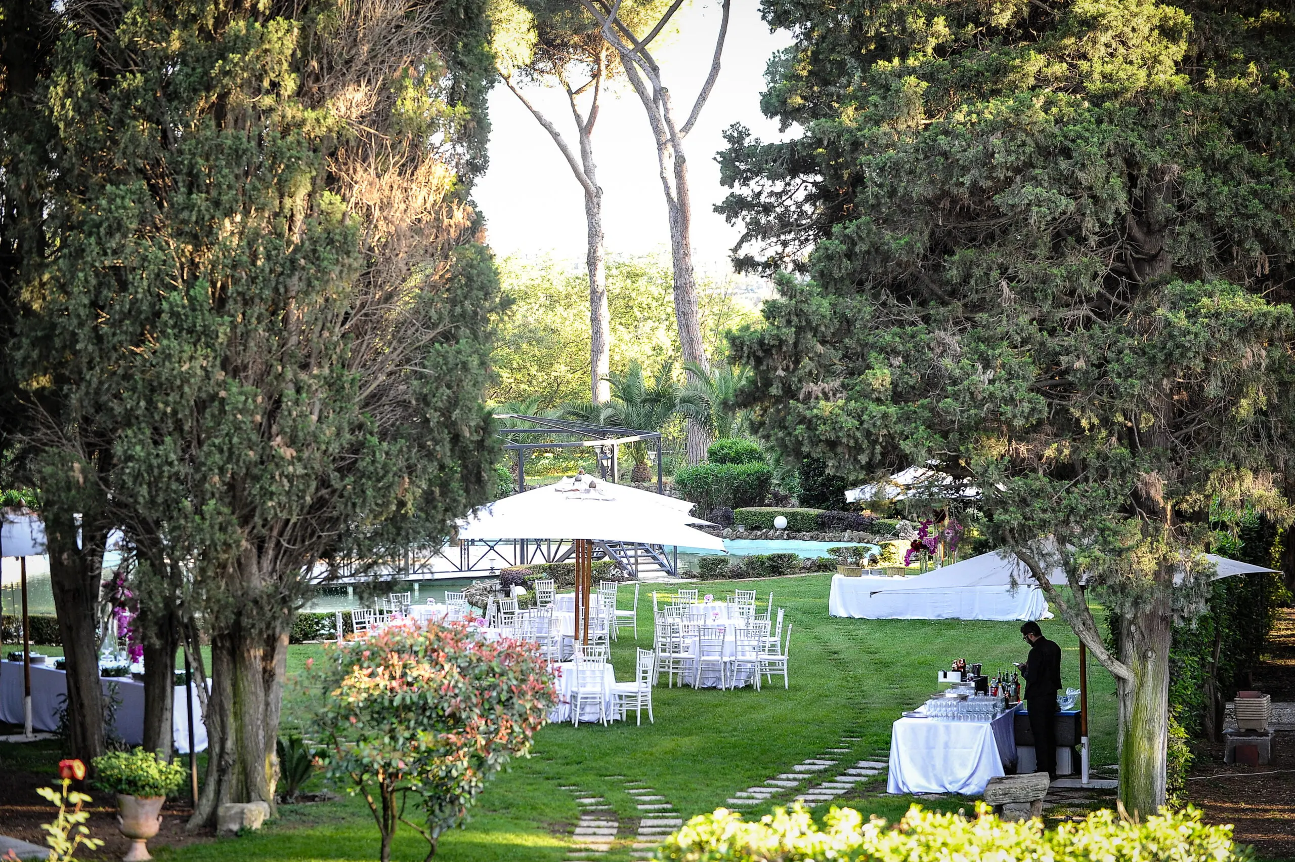 Tavoli rotondi con tovaglie bianche e sedie chiavarine allestiti sul prato del parco, fra cipressi e pini, con la piscina sullo sfondo