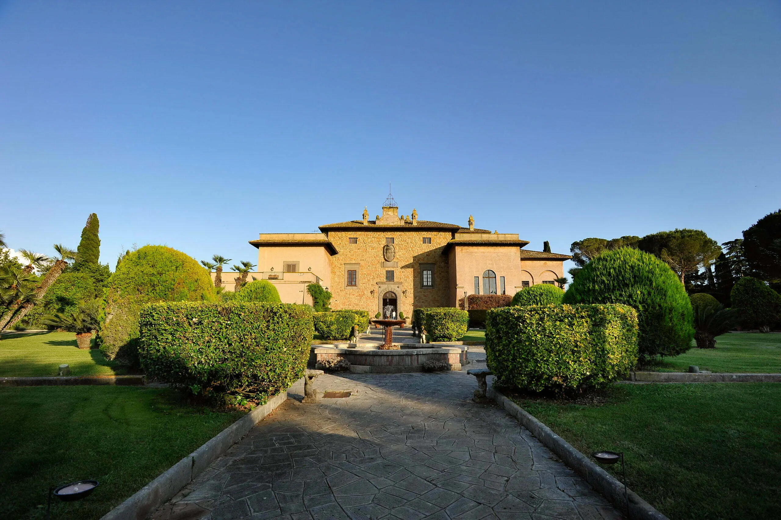 La villa vista frontalmente dal viale lastricato: facciata in blocchi di tufo dorato con torretta centrale e mascherone in pietra sopra il portale ad arco, tetto a padiglione in coppi, una fontana circolare in asse con l’ingresso, siepi potate a parallelepipedo ai lati del vialetto, cipressi e palme, cielo azzurro terso