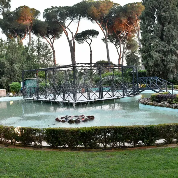 Il laghetto ornamentale con getti d’acqua a ventaglio, al centro un’isola con pergolato in ferro battuto raggiungibile da un ponticello, bordo in pietra e siepe di bosso potata tutt’intorno, alti pini marittimi e cipressi sullo sfondo