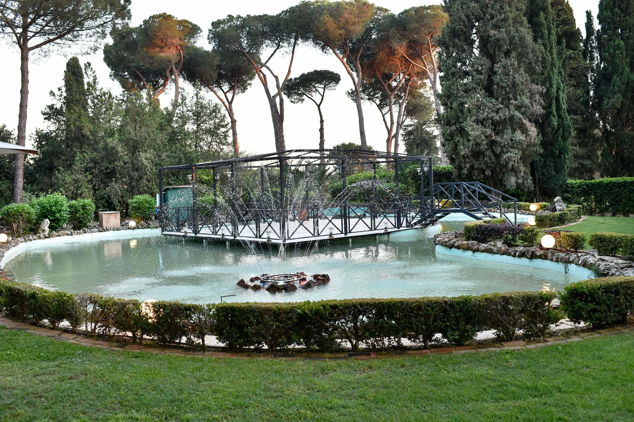 Il laghetto ornamentale con getti d’acqua a ventaglio, al centro un’isola con pergolato in ferro battuto raggiungibile da un ponticello, bordo in pietra e siepe di bosso potata tutt’intorno, alti pini marittimi e cipressi sullo sfondo